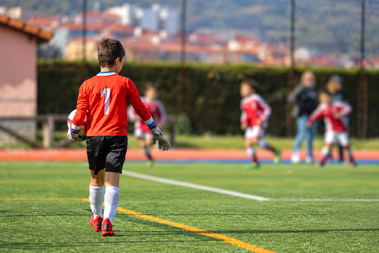 Niño portero con camiseta roja y dorsal 1 camina por el campo con el balón bajo el brazo mientras sus compañeros juegan al fondo.
