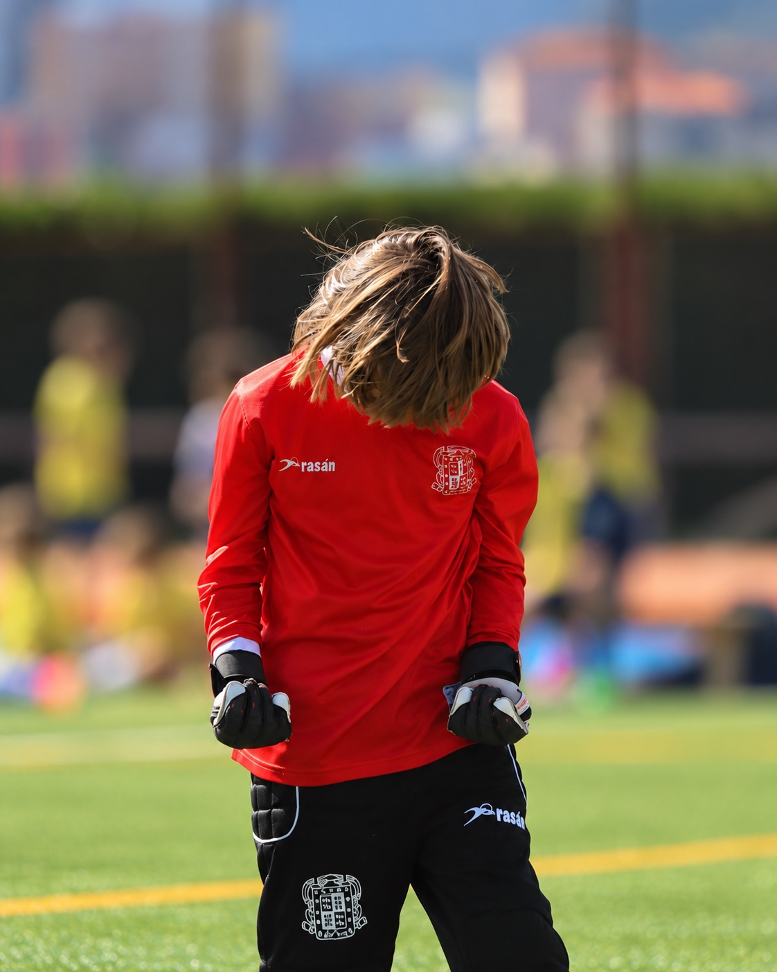Niño portero con camiseta roja y guantes celebra con los puños cerrados sobre el césped, con compañeros desenfocados al fondo.