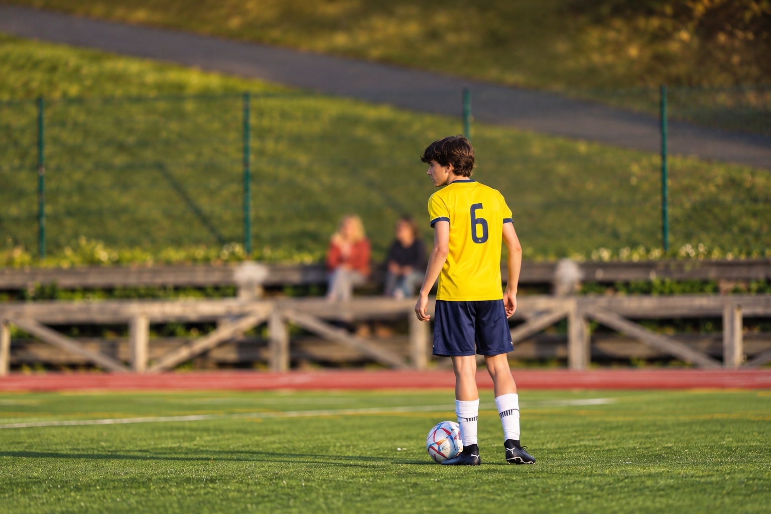 Jugador con camiseta amarilla y dorsal 6 espera junto al balón en el campo al atardecer, con dos personas mirando desde la grada.